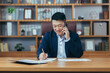 © Liubomir - Successful Asian businessman working with documents in a classic office, man talking on the phone and smiling