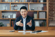 © Liubomir - Happy asian businessman working in classic office, looking at camera and smiling gesturing with hands, video call and online meeting