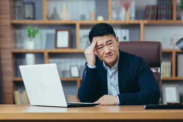 Upset businessman sitting at work in office, asian pensive, working on laptop