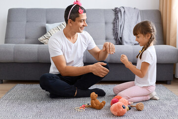 Image of smiling optimistic man wearing white t shirt and child's hair ban with flower sitting on floor and playing with his daughter, cute female kid painting father nails.