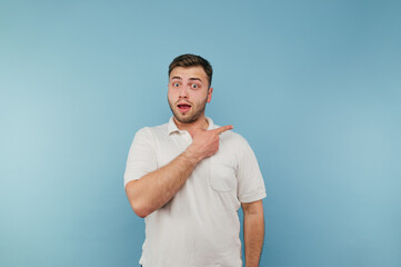 Shocked young man in a white T-shirt with bristles points his finger to the side with a surprised face on a blue background.