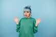 © bodnarphoto - Surprised hipster girl in green sweater with raised hands shocked looking at camera on blue background