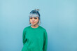 Portrait of unhappy girl in green sweatshirt and blue hair standing and looking at camera with serious face isolated on blue background.