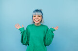 Fototapeta Uliczki - Positive teen girl with blue hair and in a green sweater rejoices with raised hands on a colored background.