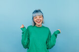 Fototapeta Uliczki - Joyful girl with blue hair in a green sweatshirt rejoices with a smile on his face and closed eyes on a blue background