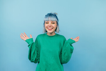 Positive teen girl with blue hair and in a green sweater rejoices with raised hands on a colored background.