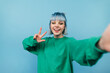 © bodnarphoto - Positive girl in a green sweater and blue hair takes a selfie with a smile on his face and shows a gesture of peace isolated on a blue background