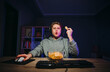 Shocked young man in a headset sits at night at the computer and eats chips from a plate with a surprised face staring at the screen.