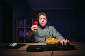 Wall Mural - Serious guy in casual clothes sits on the computer in a headset with a drink in his hands and a plate of chips, looks at the screen and plays online games