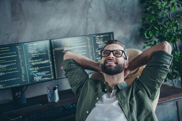 Photo of clever dreamy man wear green shirt spectacles having rest after developing software hands behind head indoors workshop workplace