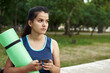 © etonastenka - Close-up of young sporty woman in blue sports clothing holding exercise mat and looking at her mobile phone in her hand while standing outdoors