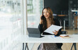 © PaeGAG - Happy smiling asian business woman working on laptop at office, using smart phone. Businesswoman sitting at her working place