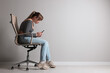 © New Africa - Young woman with poor posture using smartphone while sitting on chair near grey wall, space for text
