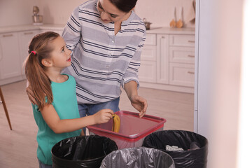 Sticker - Young woman and her daughter throwing banana peel into trash bin in kitchen. Separate waste collection