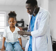 © Nassorn - African American doctor examining little girl patient heartbeat with stethoscope for proper treatment. Male nurse wears white coat performs physical checkups female kid who sitting on a hospital bed.