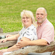 © mitarart - Portrait of happy elderly couple sitting on bench in park. Smiling old people looking at camera. Summer day in nature. Green fields in background.