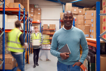 Portrait Of Manager With Digital Tablet In Busy Modern Warehouse With Staff In Background