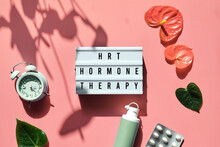 Text HRT Replacement Therapy On Light Box. Menopause, Hormone Therapy Concept. Oestrogen Replacement Therapy Awareness. Pink Background With Alarm Clock, Exotic Leaves, Pills, Estrogene Gel.