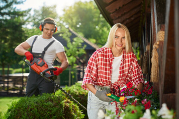 Wall Mural - Strong caucasian man in uniform using electric trimmer for pruning bushes at garden while charming woman cutting dry leaves on flower in pots. Seasonal work outdoors. 