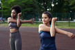 © etonastenka - Two fit smiling young woman working out together in a park doing stretching exercise and hand extensions to improve suppleness