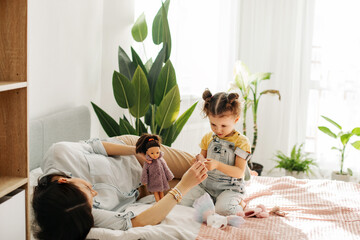 A charming little girl plays with dolls with her mom on the bed during the day. Motherhood, caring, time together