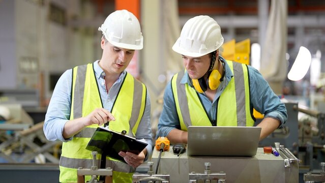 Two specialist engineer team with safety helmet and vast uniform using paper chart and laptop consulting together enjoying work time working in steel and metal manufacturing plant
