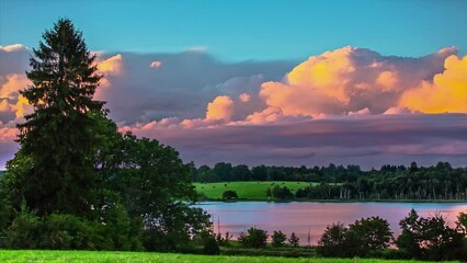 Wall Mural - Timelapse footage of fast moving white puffy clouds on blue clear sky over rural landscape with greenlands and lake surrounded by trees.