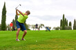 © trattieritratti - Golfer on a golf course in summer on a day with clouds, hitting the ball with a golf club in the direction of the flag.