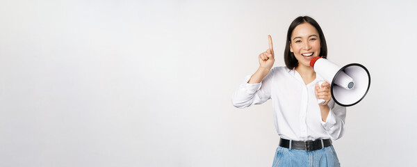 Wall Mural - Smiling happy asian girl talking in megaphone and pointing up, announcing discount promo, showing advertisement on top, standing over white background