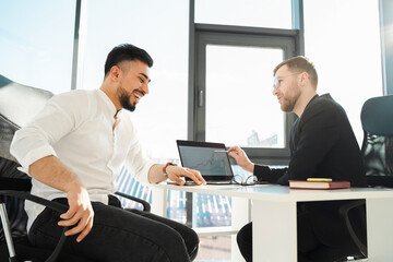 Two laughing businessmen in the office at the laptop analyze the growth of the company