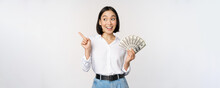 Smiling Young Modern Asian Woman, Pointing At Banner Advertisement, Holding Cash Money Dollars, Standing Over White Background
