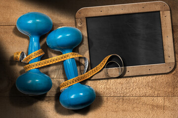 Close-up of two old blue dumbbells for workout or fitness (free weights) with orange tape measure and empty blackboard with copy space, on a wooden background.