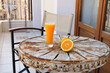 Orange juice in a tall transparent glass on an old metal table with beautiful patterns. Next to it is a fresh orange cut in half. A small metal table with chairs on the balcony.