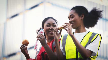 smile two African american woman foreman worker  or woman maintenance engineer in reflective vest safety jacket sits down on old truck relaxing, eats bread snacks and water during brunch break