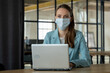 © Grustock - Woman in the office is working in a medical face mask behind a laptop.