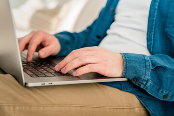 Canvas Print - hands a young man in a blue shirt using a laptop on his lap while sitting on the couch. cozy conditi