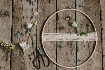 Making stylish spring wreath with beautiful fresh flowers. Wooden hoop, thread, scissors, and flowers on rustic wooden background flat lay. Modern and creative floral handmade decor