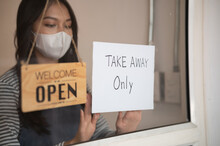 Young Asian Woman Restaurant Or Coffee Shop Owner Wear Face Mask Attaching Take Away Only Sign On Front Door, Entrepreneur Opening Cafe Only For Take Away Orders During Coronavirus Pandemic.