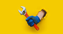 A Right Hand In A Blue Knitted Glove Holds A Red Monkey Wrench. Torn Hole In Bright Yellow Paper. The Concept Of A Worker, Labor Migrant, A Master Of His Craft. Copy Space.