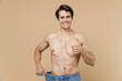 Satisfied young man 20s with naked torso show loose pants show thumb up isolated on pastel pastel beige color background Body care healthcare fitness sport bodybuilding diet loosing weight concept.