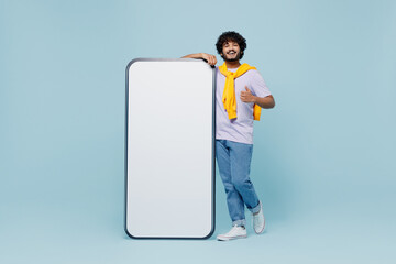 Wall Mural - Full size jubilant fun young bearded Indian man 20s wear white t-shirt stand near big mobile cell phone with blank screen workspace area isolated on plain pastel light blue background studio portrait