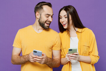 Young smiling nody curious couple two friends family man woman together in yellow casual clothes hold use mobile cell phone look to each other smartphone isolated on plain violet background studio.