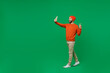 Full body young fun smiling happy caucasian man 20s in orange sweatshirt hat doing selfie shot on mobile cell phone post photo on social network show v-sign isolated on plain green background studio