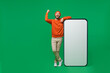 Full body young happy man 20s in orange sweatshirt hat stand near big blank screen mobile cell phone with workspace copy space mockup area do winner gesture isolated on plain green background studio