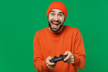 Sticker - Young overjoyed smiling happy caucasian man 20s wear orange sweatshirt hat hold in hand play pc game with joystick console isolated on plain green background studio portrait. People lifestyle concept.