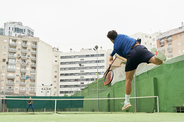 Young male tennis player in blue sportswear jumps and hits serve with ball and racket in tennis match