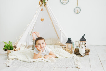 Little caucasian boy laughing while lying in a toy kids teepee in a white scandinavian christmas interior.