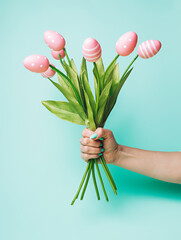 Wall Mural - Woman's hand holding bouquet of tulip flowers with colored eggs instead of buds on pastel green background. Creative Easter holiday concept. Natural traditional holiday greeting card. Spring bloom.