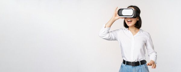 Wall Mural - Excited young woman enter virtual reality in her glasses. Asian girl using vr headset, standing over white background