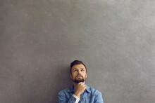 Serious Handsome Man In Modern Casual Denim Jacket Leaning On Wall, Thinking And Looking Up At Copy Space Above Head. Young Guy Dreaming About Something Standing On Grey Text Space Background